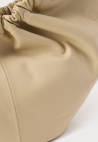 Μπεζ δερμάτινη τσάντα χειρός με σούρα στο πάνω μέρος και ορατή ραφή, με μικρό χρυσό λογότυπο "Coccinelle" κοντά στο κάτω δεξιά μέρος.