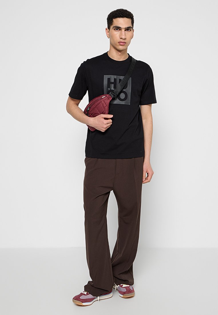 T-shirt noir à manches courtes avec un logo, pantalon large marron et sac à bandoulière bordeaux. Les chaussures présentent des accents rouges sur une base grise.