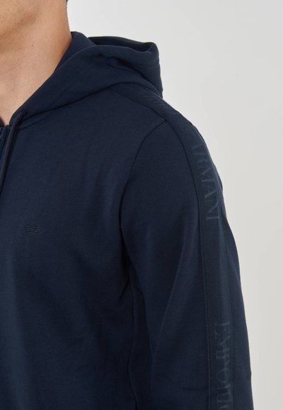 Mörkblå bomullshuvtröja med dragsko i huvan, med en liten logotyp på bröstet och ton-i-ton text längs ärmarna.