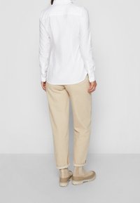 Hvid langærmet skjorte med knaplukning ved manchetterne, parret med beige, løstsiddende bukser og brune ankelstøvler. Enkel design, glat stoftekstur.