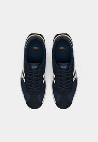 Ténis azul-marinho feitos de tecido texturado, com riscas brancas e detalhes pretos. Incluem atilhos redondos e um logótipo na língua.