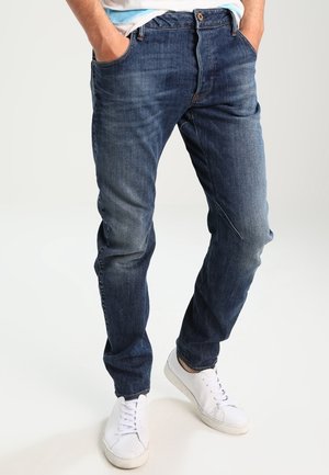 Dunkelblaue Jeans aus Denim mit geradem Bein, ausgestattet mit Fronttaschen und einem Reißverschluss. Getragen mit weißen Sneakers.