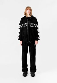 Chaqueta con capucha negra con texto gráfico blanco y rayas, combinada con pantalones negros y zapatos negros texturizados. Diseño sencillo y casual.