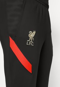 Zwarte sportbroek met een rode diagonale streep en een gouden geborduurd Liverpool Football Club-logo aan de linkerkant.