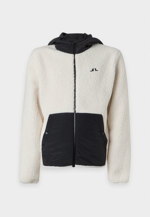 Homme grand portant une veste polaire blanche et noire zippée avec capuche, un pantalon noir et des baskets noires sur un fond blanc uni.