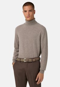 Pull col roulé marron clair en tissu doux, associé à une ceinture tressée dans un ton vert-brun atténué. Un pantalon marron vient parfaire la tenue.