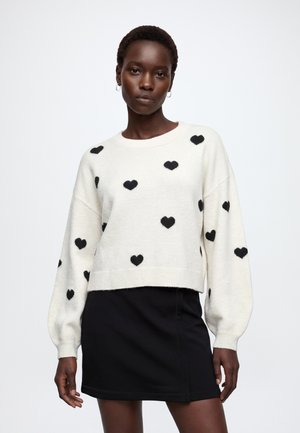 Kvinde iført en hvid sweater med sorte hjerter og et sort skørt, stående mod en ensfarvet lys baggrund.