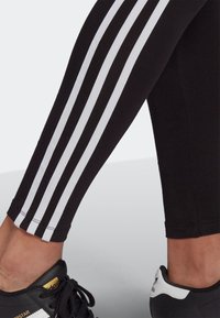 Black leggings with three vertical white stripes along the side, fitted design, and a smooth texture. Paired with black sneakers.