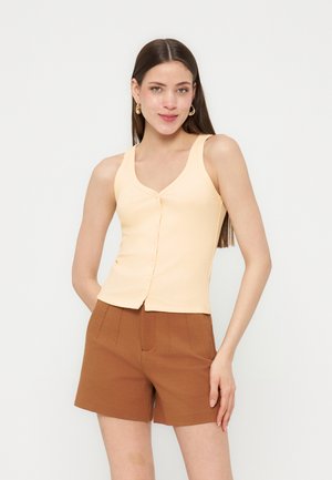 Młoda kobieta o długich brązowych włosach, nosząca złote kolczyki koła, beżową bezrękawnikową bluzkę na guziki oraz brązowe szorty z wysokim stanem, pozująca na białym tle.