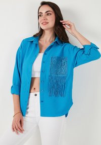 Camicia blu strutturata con tasca in pizzo e dettagli frangiati. Maniche arrotolate e vestibilità comoda. Indossata sopra un crop top bianco.
