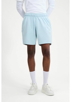Model trägt hellblaue elastische Shorts mit hohem Bund, weiße Socken und weiße Sneakers und steht vor einem schlichten weißen Hintergrund.