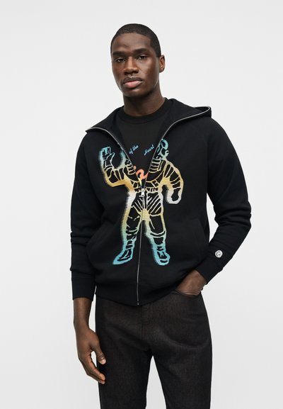 Άνδρας που φοράει μαύρο φούτερ με φερμουάρ και πολύχρωμο σχέδιο αστροναύτη, μαζί με μαύρο παντελόνι, στέκεται μπροστά σε απλό φωτεινό φόντο.