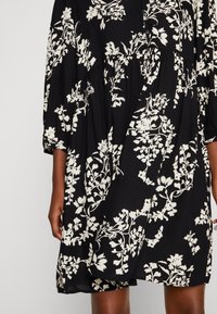 Robe noire avec un motif floral blanc, manches courtes, col rond et taille froncée. Fabriquée à partir d'un tissu léger avec une texture fluide.