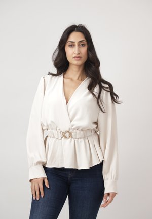 Donna con capelli lunghi e scuri che indossa una camicetta bianca con cintura e jeans blu scuro, in piedi davanti a uno sfondo chiaro e uniforme.