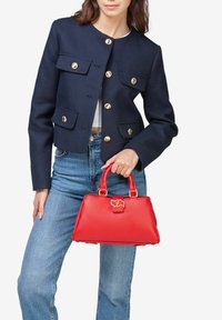 Bolso de mano de cuero rojo con forma estructurada, acentos de hardware dorado, un asa superior y un detalle de cierre. Lleva puesto con una chaqueta azul marino y jeans.