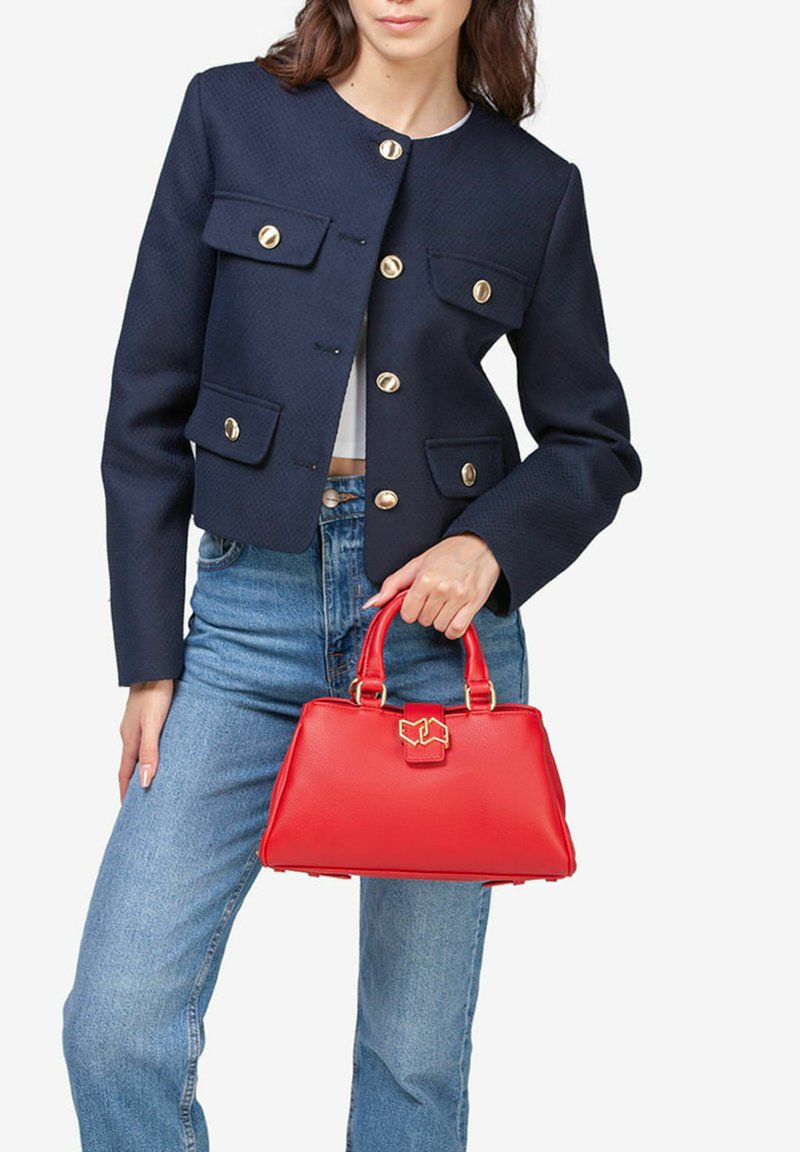 Bolso de mano de cuero rojo con forma estructurada, acentos de hardware dorado, un asa superior y un detalle de cierre. Lleva puesto con una chaqueta azul marino y jeans.