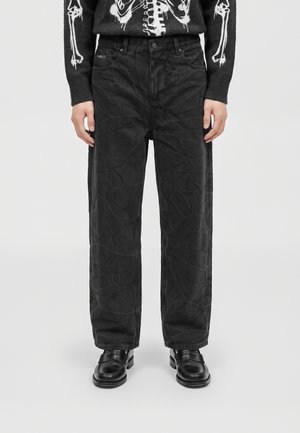 Modèle portant un jean noir ample à motif craquelé, un pull à imprimé squelette et des chaussures en cuir noires sur un fond uni.