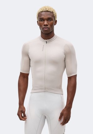 Homme athlétique aux cheveux blonds courts portant un maillot de cyclisme beige ajusté à manches courtes et un cuissard rembourré gris clair, face à l'avant.