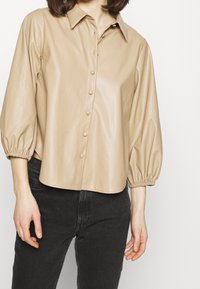 Chemise beige en simili cuir avec un col pointu, des boutons sur le devant et des manches trois-quarts bouffantes. Texture lisse, coupe légèrement courte.