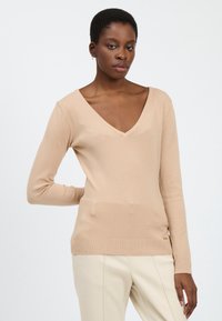 Beige V-Ausschnitt-Pullover mit langen Ärmeln, aus weichem, strukturiertem Stoff. Gerippter Saum und Bündchen, mit einem subtilen Logodetail an der Seite.