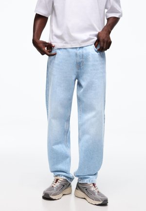 Person trägt lockere hellblaue Jeans, weißes T-Shirt und graue Sneakers, steht mit den Händen in den Taschen vor weißem Hintergrund.