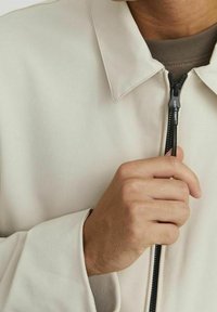 Giacca cremoso chiaro con zip, collo alla coreana, tessuto liscio e zip nera a contrasto, che presenta un design minimalista.