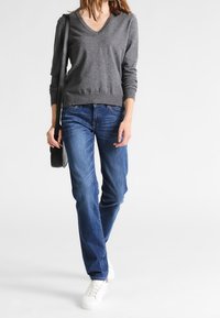 Grau-gestreifter V-Ausschnitt-Pullover, kombiniert mit blauen Jeans. Das Model trägt weiße Sneakers und trägt eine schwarze Umhängetasche. Weiches Material, lässige Passform.