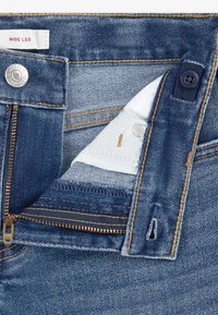 Traper hlače širokih nogavica plave boje, s patentnim zatvaračem, gumbima kao ukrasom i mekanom tkaninom postavom unutar pojasa.