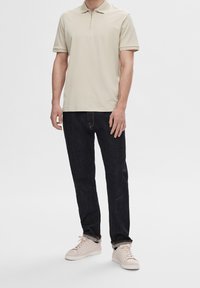 Polo beige à col zippé avec manches courtes, associé à un jean bleu marine et des baskets beige clair. Design simple, texture lisse.