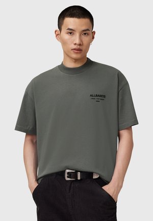 T-shirt à manches courtes vert foncé en coupe décontractée avec un col rond, arborant un logo noir brodé sur la poitrine gauche. Porté avec un pantalon en velours côtelé noir.