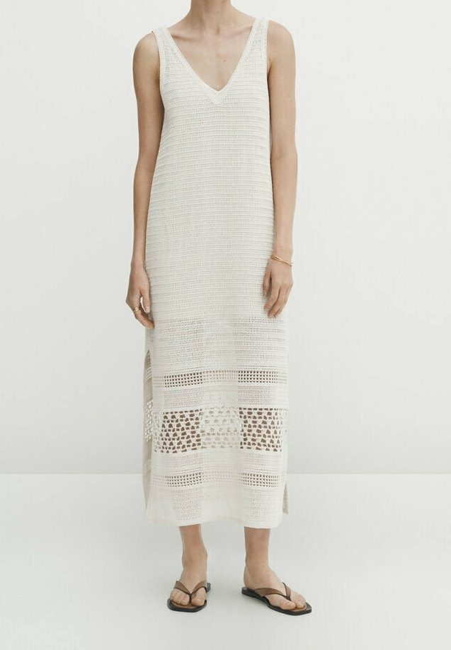 Robe midi blanche sans manches en crochet avec encolure en V et fentes latérales, portée avec des sandales plates noires à lanières sur une personne debout.