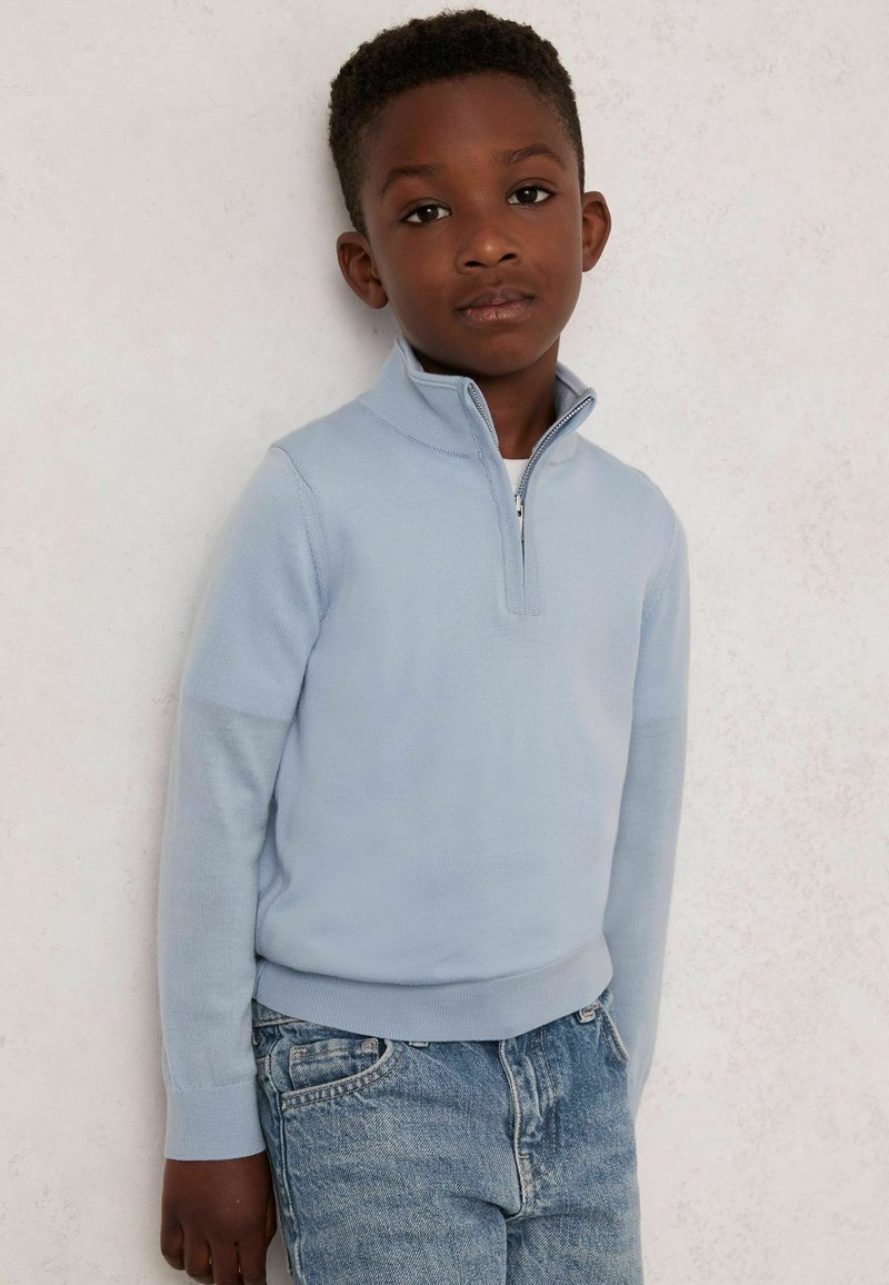 Hellblaues Zip-Up-Sweatshirt mit langen Ärmeln, gerippten Bündchen und Saum, kombiniert mit hellblauen Jeans, vor einem neutralen Textur-Hintergrund.