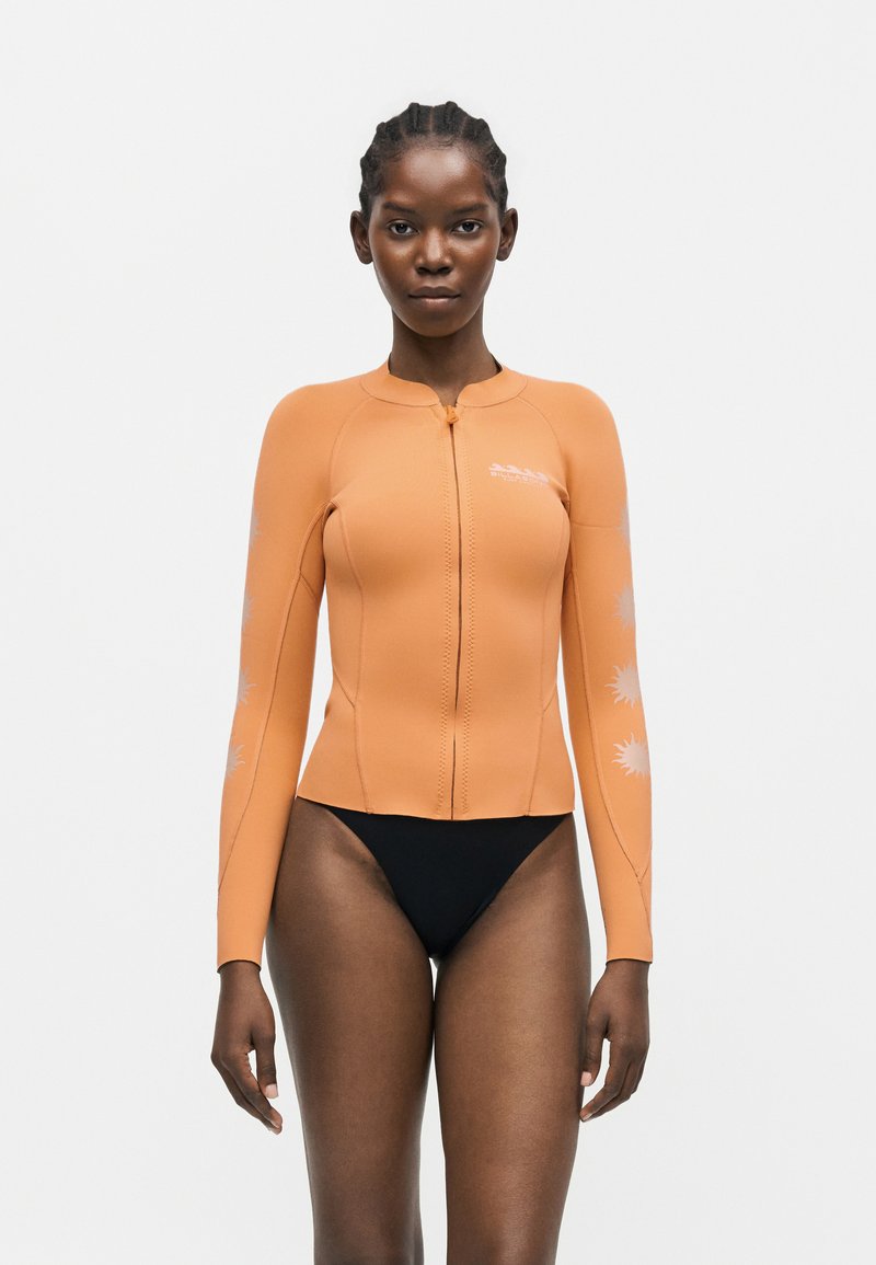 Λεπτή γυναίκα με πλεγμένα μαλλιά που φοράει εφαρμοστό πορτοκαλί μπουφάν καταδυτικού κοστουμιού με φερμουάρ και μαύρο μπικίνι, στέκεται μπροστά από ένα απλό φόντο.