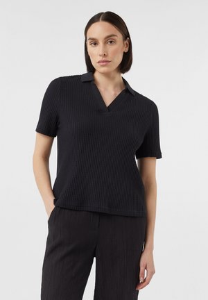 Schwarzes Poloshirt mit kurzen Ärmeln aus strukturiertem Waffelstrick, mit V-Ausschnitt und kontrastierendem Kragen. Getragen mit dunklen, strukturierten Hosen.