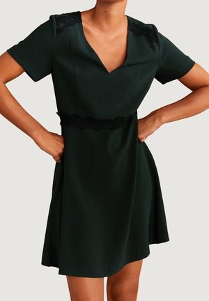 Schwarzes, knielanges Kleid mit kurzen Ärmeln, V-Ausschnitt und Spitzenakzenten an Schultern und Taille, getragen von einer Person mit den Händen in der Hüfte.