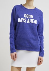 Persona vistiendo un sudadera azul con el texto "BUENOS DÍAS POR DELANTE" y una falda blanca, de pie contra un fondo claro y liso.