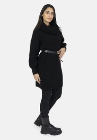 Robe pull oversized noire en tricot avec un col drapé, cintrée à la taille par une ceinture noire dotée d'une boucle en forme de cœur. Portée avec des leggings noirs et des bottes de combat.