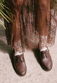 Zapatos de cuero marrón con cordones prominentes, punta redondeada y suela acolchada, combinados con calcetines blancos, debajo de una falda de encaje marrón semitransparente.