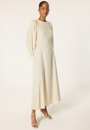 Femme portant une longue robe crème avec des manches longues bouffantes et des sandales à talons hauts avec bride à la cheville, debout devant un fond clair uni.