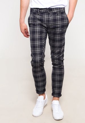 Mann trägt eng geschnittene marineblaue und graue karierte Hose mit aufgekrempelten Säumen und weißen Sneakers, steht vor einfarbigem Hintergrund.