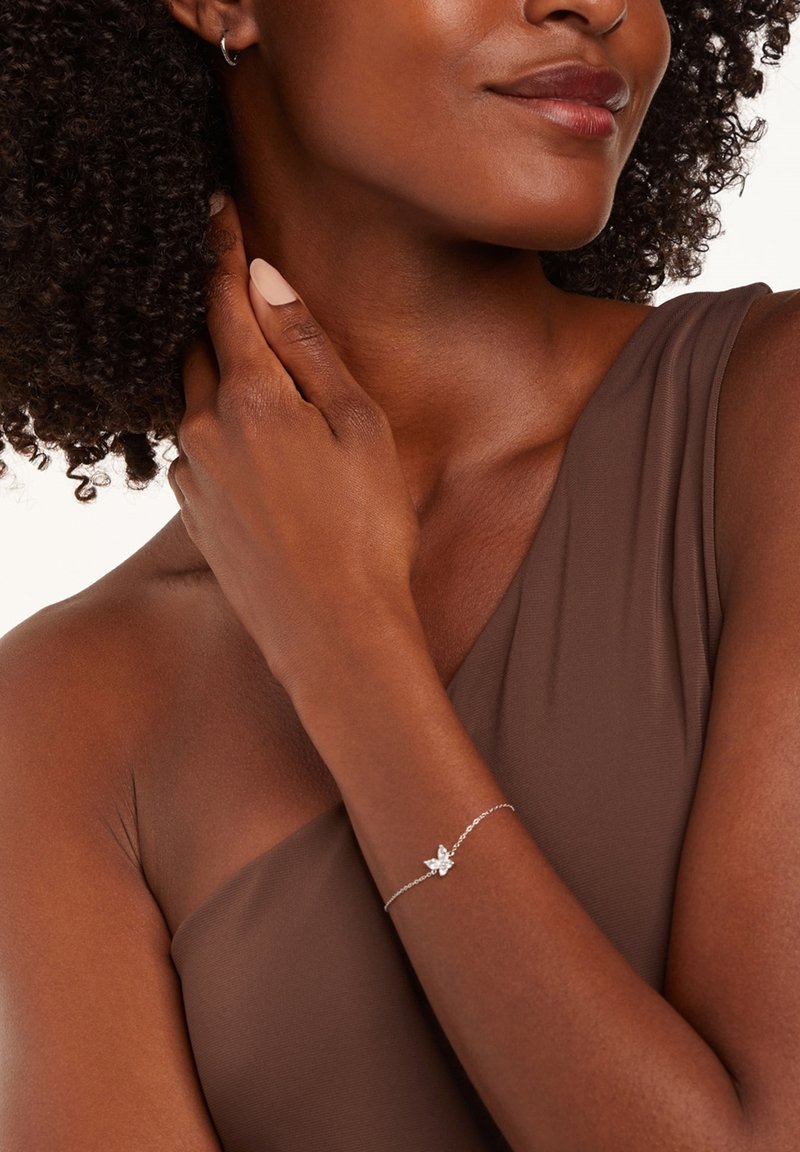 Bracelet en argent avec un motif floral, mettant en valeur une pierre claire, porté sur un bras à la peau brune contre un haut sans manches marron.