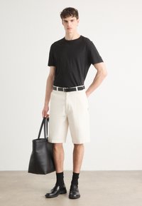 Schwarzes Rundhalsausschnitt-T-Shirt, cremefarbene Shorts, schwarzer Gürtel, schwarze Ledertasche, schwarze Slipper und schwarze Socken an einem Modell vor einem hellen Hintergrund.