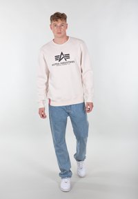 Felpa rosa chiaro con logo e testo neri, abbinata a jeans blu chiaro e sneakers bianche. Design pulito, vestibilità regolare, stile casual.