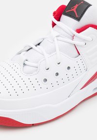 Białe buty sportowe z czerwonymi akcentami, teksturowana syntetyczna cholewka, perforowany wzór, płaskie sznurówki i gumowa podeszwa. Na języku znajduje się logo Jumpman.