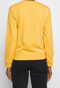 Geel sweatshirt met lange mouwen, ronde hals, ribgebreide manchetten en zoom, gladde textuur, gedragen met zwarte broek, met een eenvoudig ontwerp.