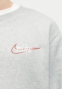 Ανοιχτό γκρι φούτερ με λευκή επιγραφή "nike" και κόκκινο λογότυπο Nike κεντημένο στο αριστερό μέρος του στήθους.