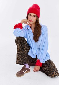 Kobieta w czerwonej czapce, niebieskiej koszuli z czerwonymi mankietami, spodniach w panterkę i brązowych sneakersach, siedząca na podłodze na białym tle.