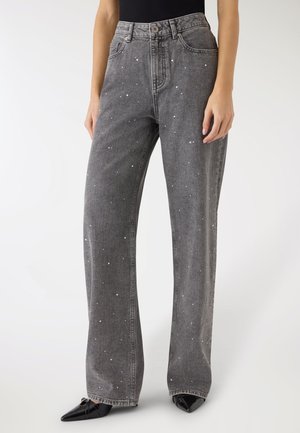 Kobieta ubrana w szare dżinsy z wysokim stanem i szerokimi nogawkami, z rozrzuconymi małymi białymi kropkami oraz czarne szpiczaste buty, stojąca na białym tle.