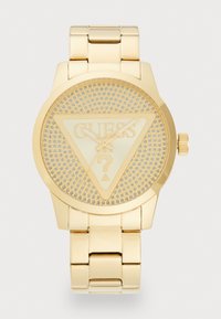 Orologio da polso in tono oro con quadrante triangolare, texture a rilievo e logo "GUESS" inciso. Presenta un bracciale metallico abbinato e una corona.