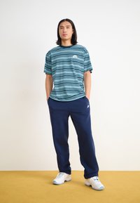 Ριγέ κοντομάνικο μπλουζάκι σε τιρκουάζ και ναυτικό μπλε, συνδυασμένο με ναυτικές φόρμες. Η μπλούζα διαθέτει μικρό λογότυπο, ενώ οι φόρμες έχουν πλαϊνές τσέπες.